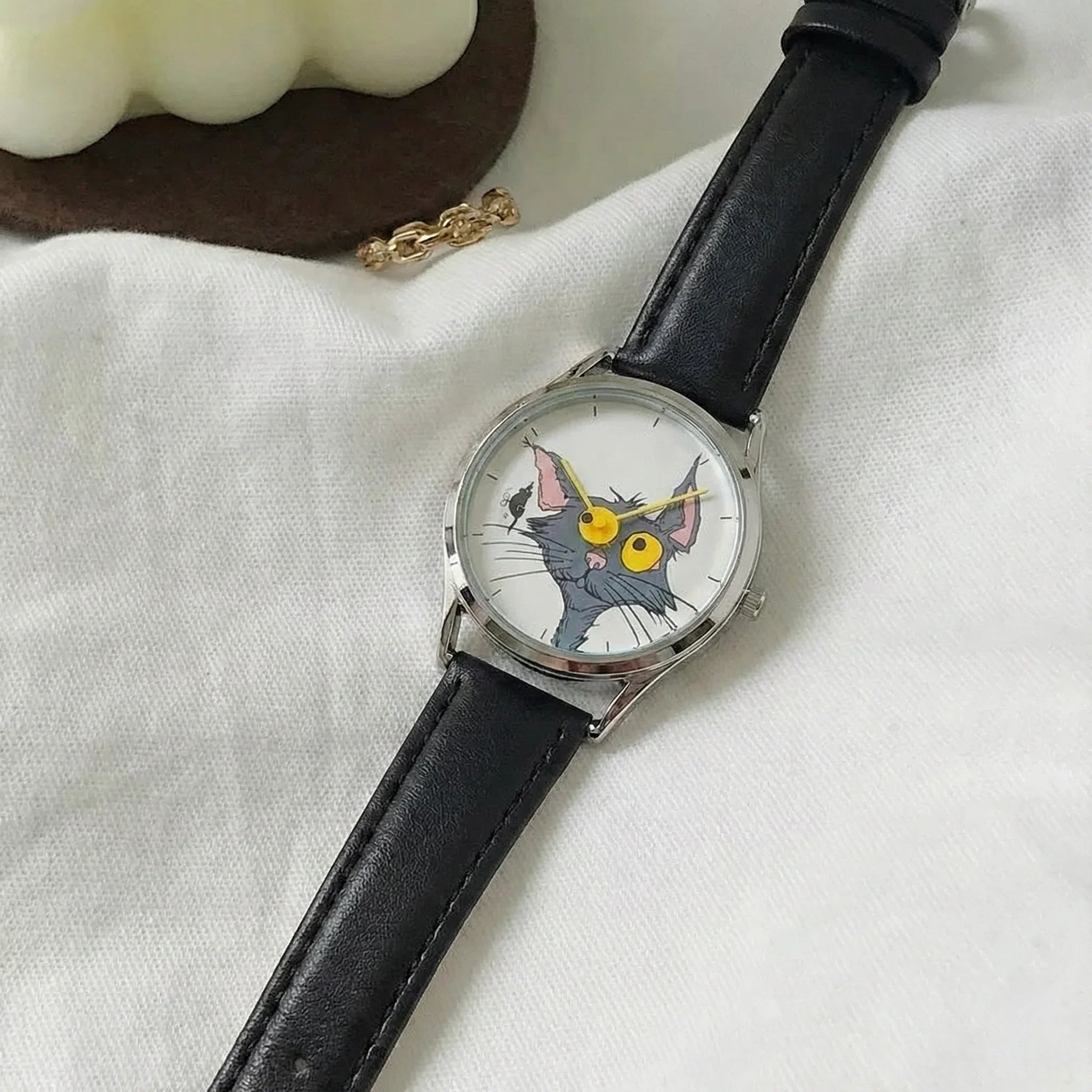 Wristwatch with a cat face design on the dial, black leather strap, on a white fabric background.